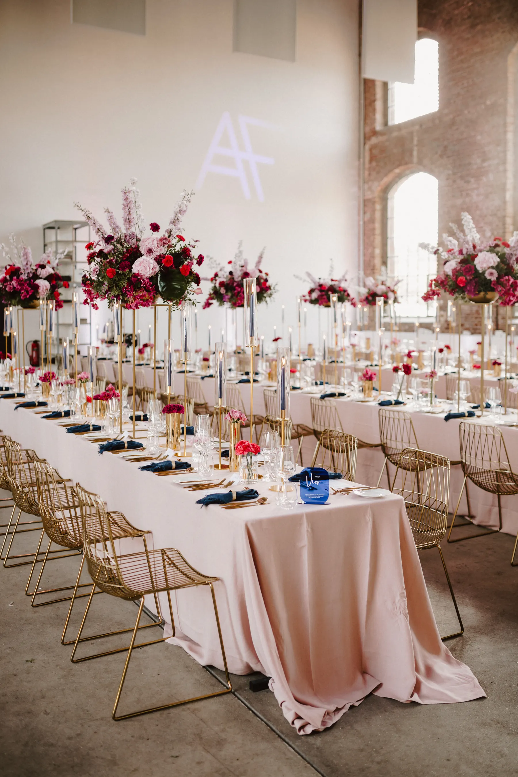 Lange Hochzeitstafeln in einer Industriehalle mit pinker Floristik und goldenen Details