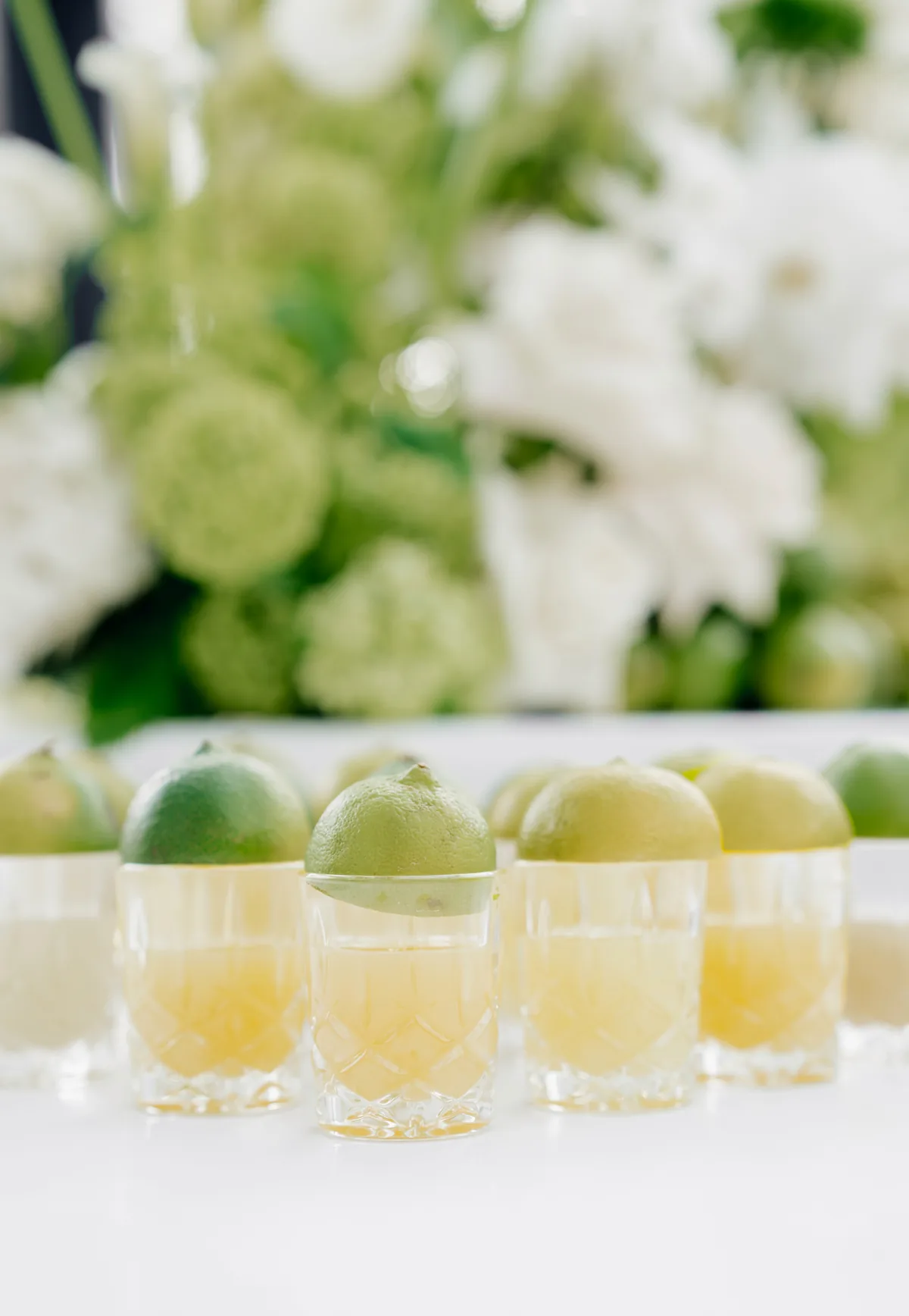 Limettenshots in Kristallgläsern vor weißer Floristik