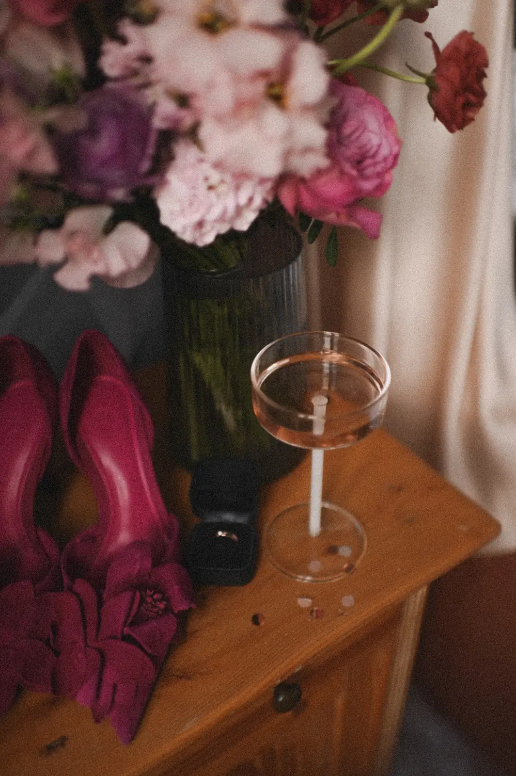 Pinke Schuhe mit Schleife, Ringbox aus Samt und ein Glas Rosé vor einem Strauß in Pink und Lila