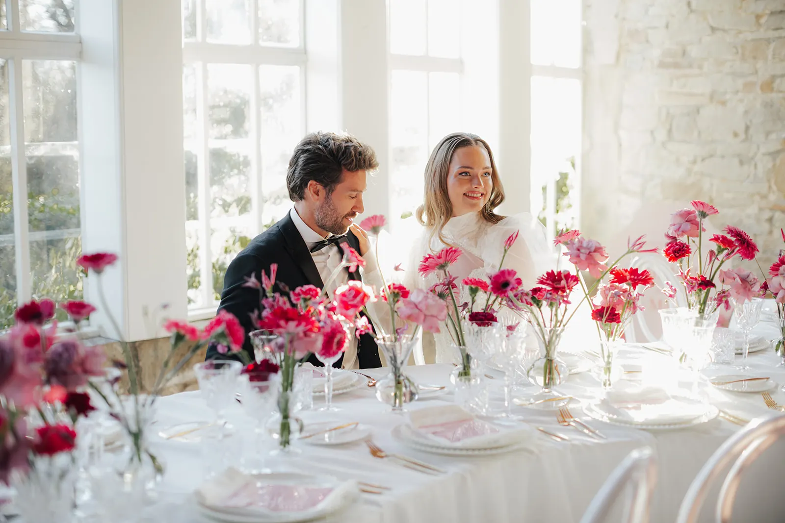 Das Brautpaar sitzt lachend an der Tafel zwischen Blumen in Pink und Rot