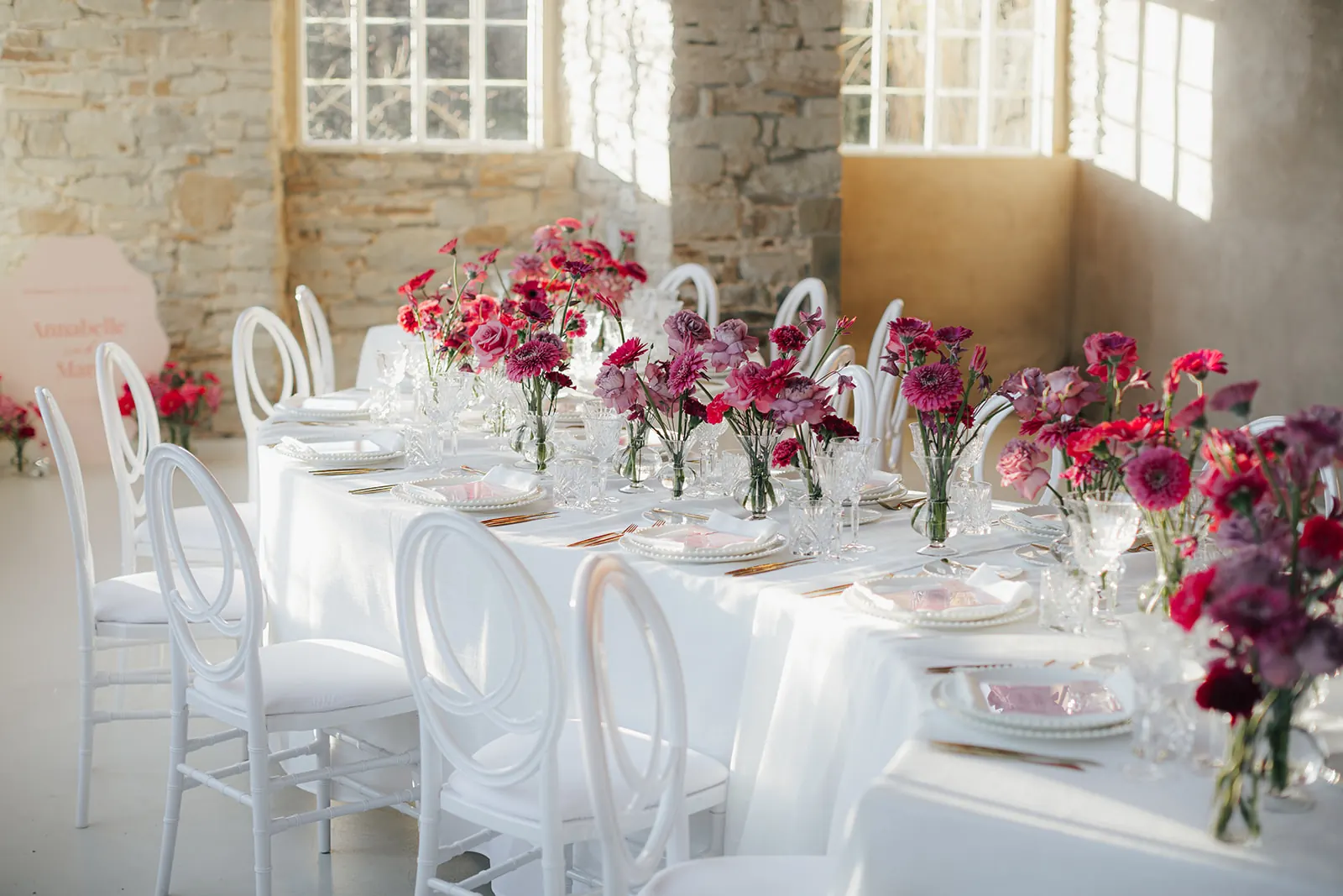 Lange Hochzeitstafel mit weißen Stühlen, Blumen in Pink, Rot und Beere und Menükarten in Rosé